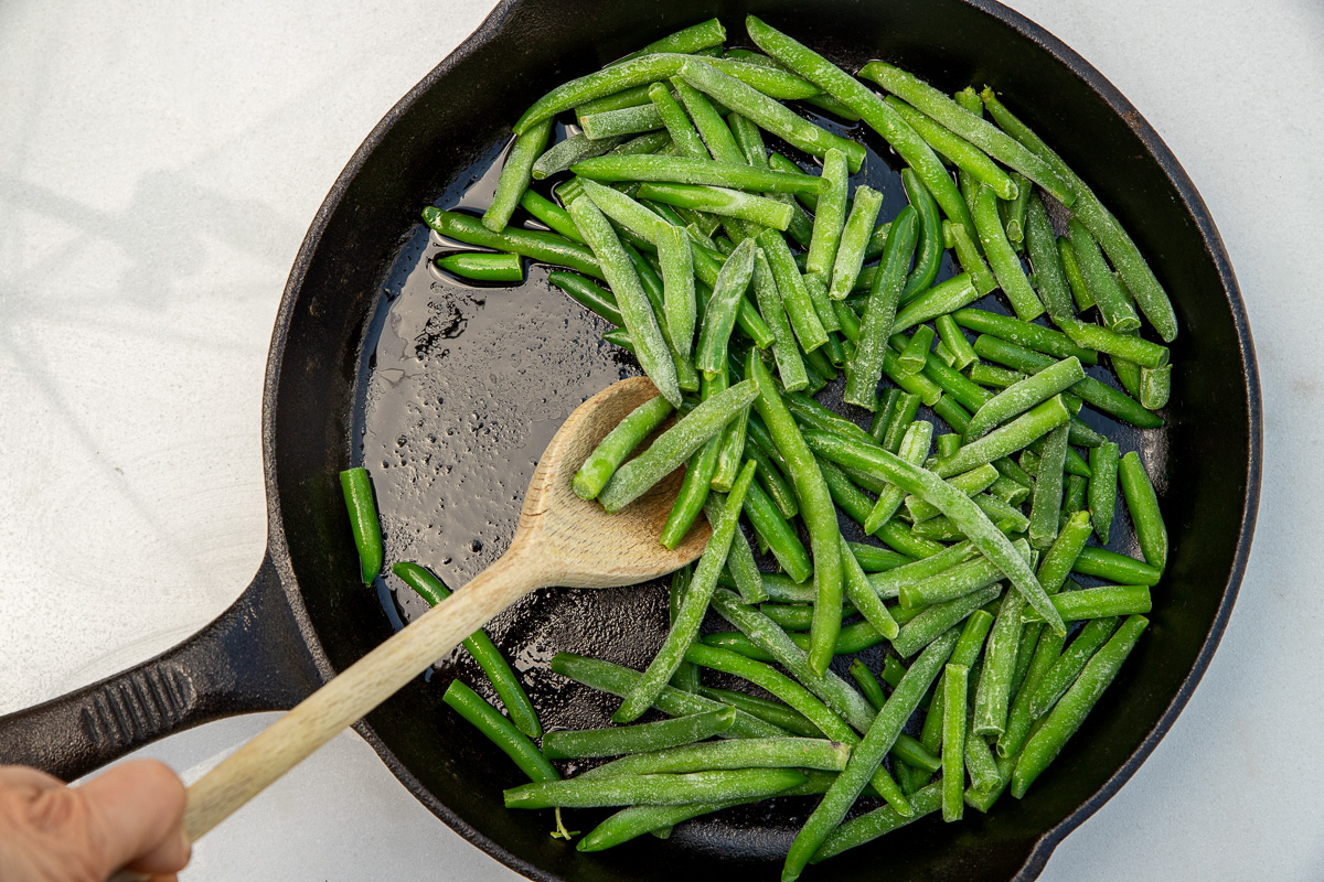 An overhead view of frozen green beans ready to be stirred with a wooden spoon in a black cast iron pan on a marble background.