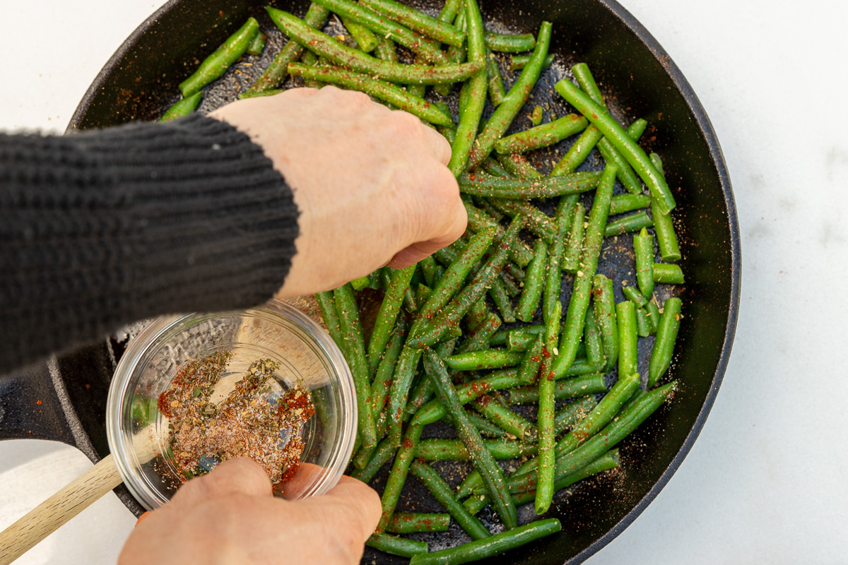 An overhead view of someone's hand adding dried mixed seasonings to a pan of frozen green beans.