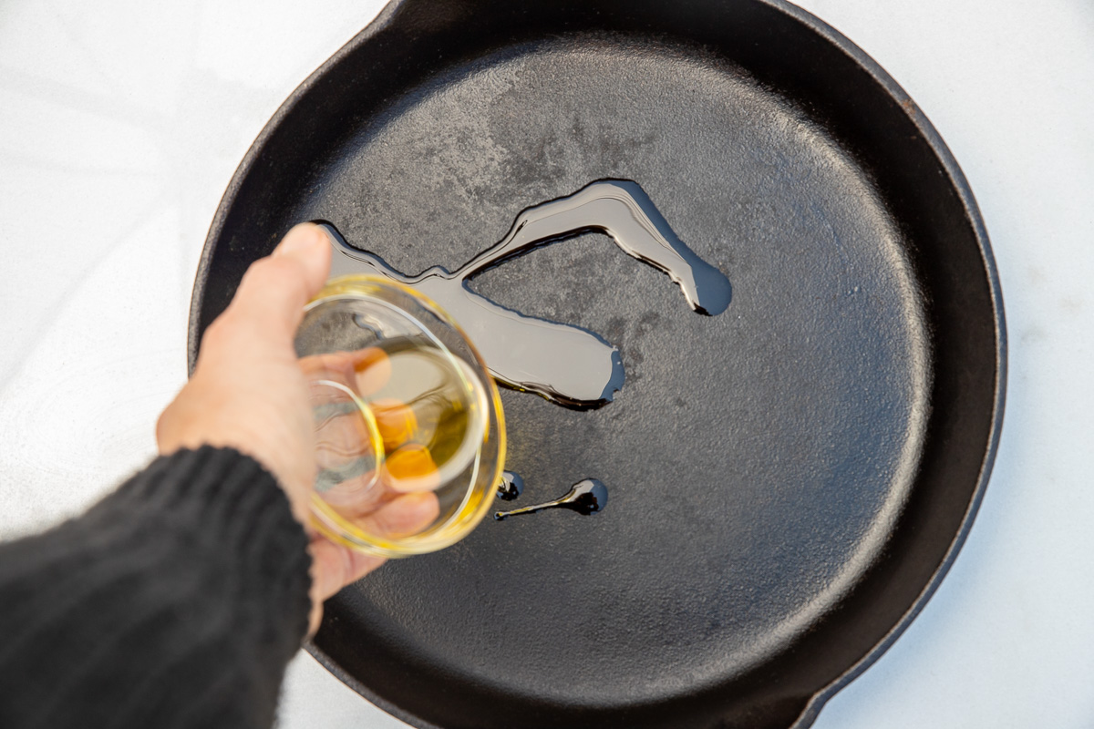 Overhead view of someone's hand pouring olive oil into a black cast iron pan from a small glass bowl.