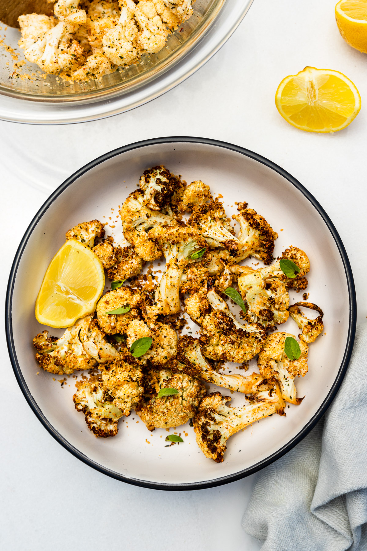 A dish of air fried cauliflower from above with fresh herbs scattered on top and a segment of lemon, with more lemon and a pale blue tea towel.