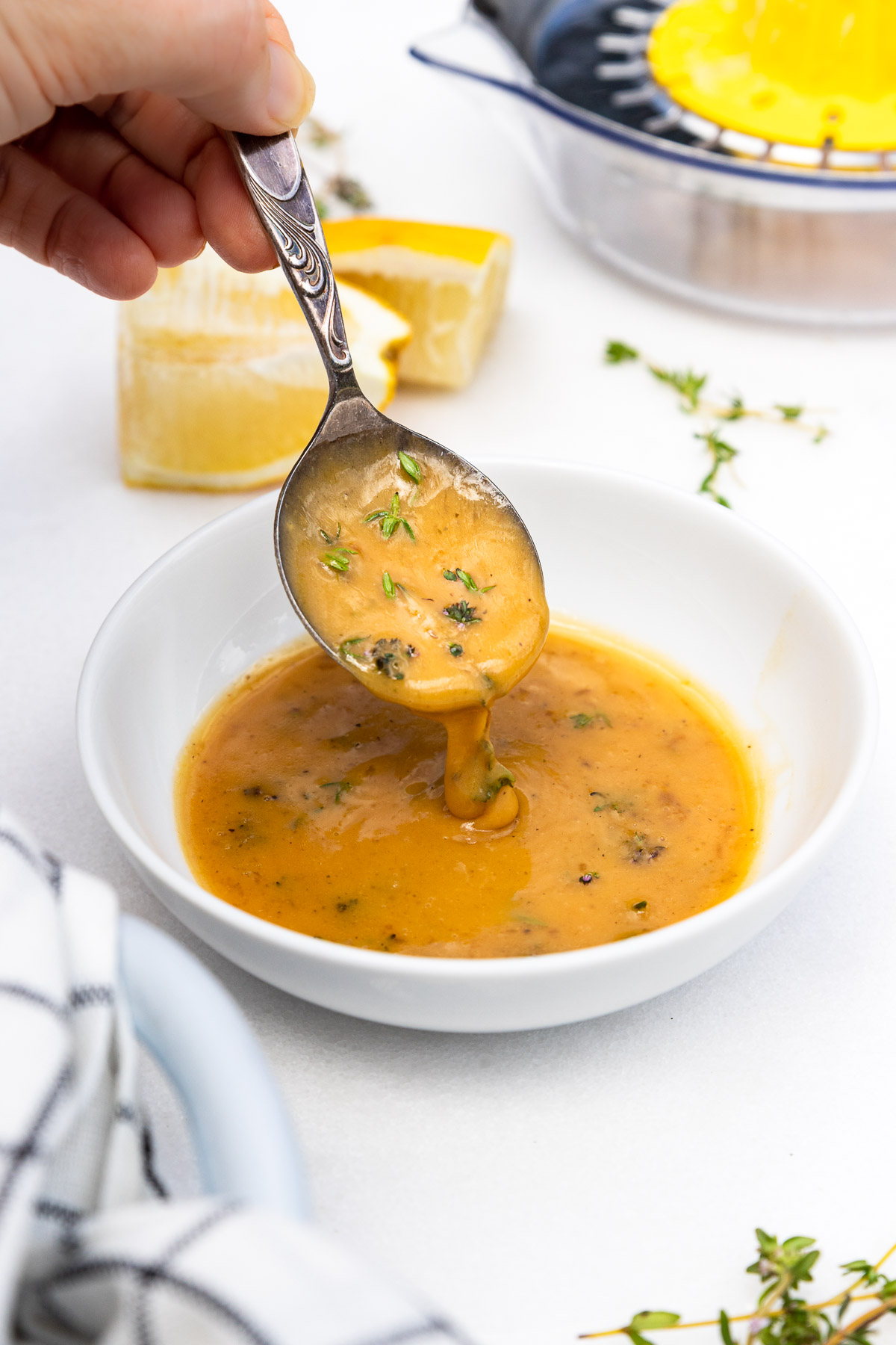 Closeup of someone's hand drizzling thick lemon, butter and herb sauce into a white dish of sauce on a marble background with checked tea towel, fresh thyme and lemons scattered around the sides.