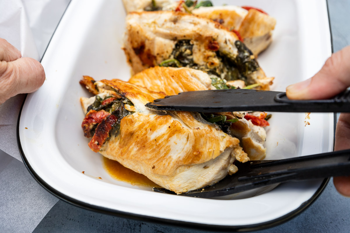 Someone with black kitchen tongs placing cooked spinach feta stuffed chicken into a white baking dish to let them rest.