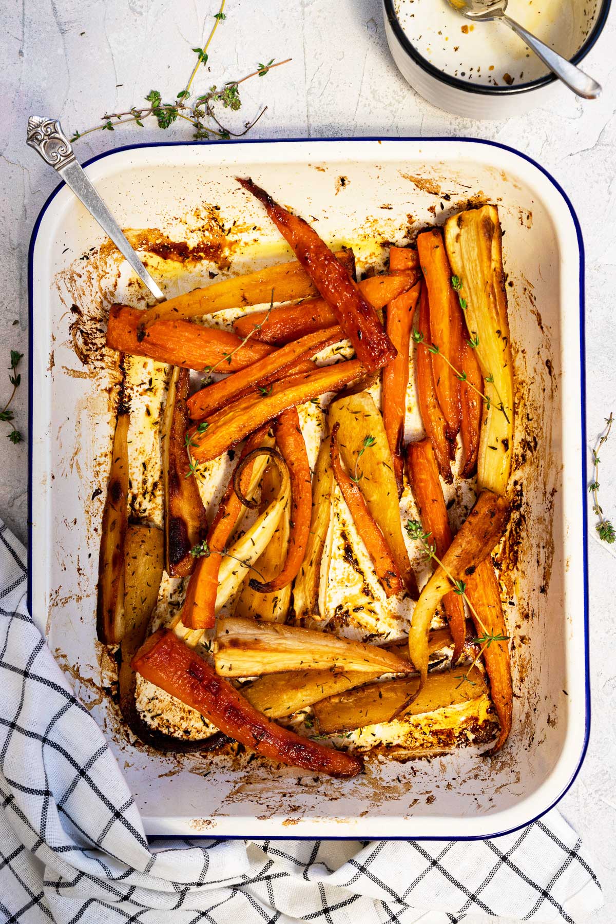 Honey roasted carrots and parsnips with thyme photographed overhead with a black and white checked tea towel in front and fresh thyme at the top, all on a textured white backgrounde.