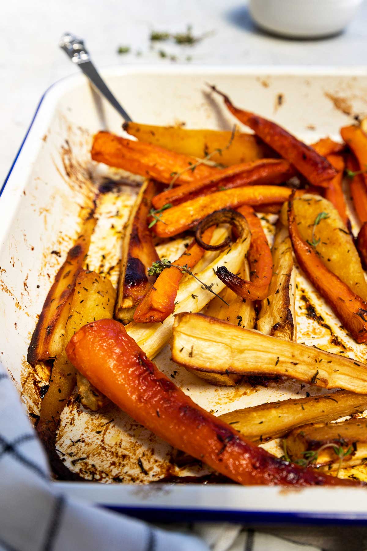 Closeup view of honey roasted carrots and parsnips in a white baking dish with a blue rim sprinkled with fresh thyme and with a blue and white checked tea towel in the foreground.
