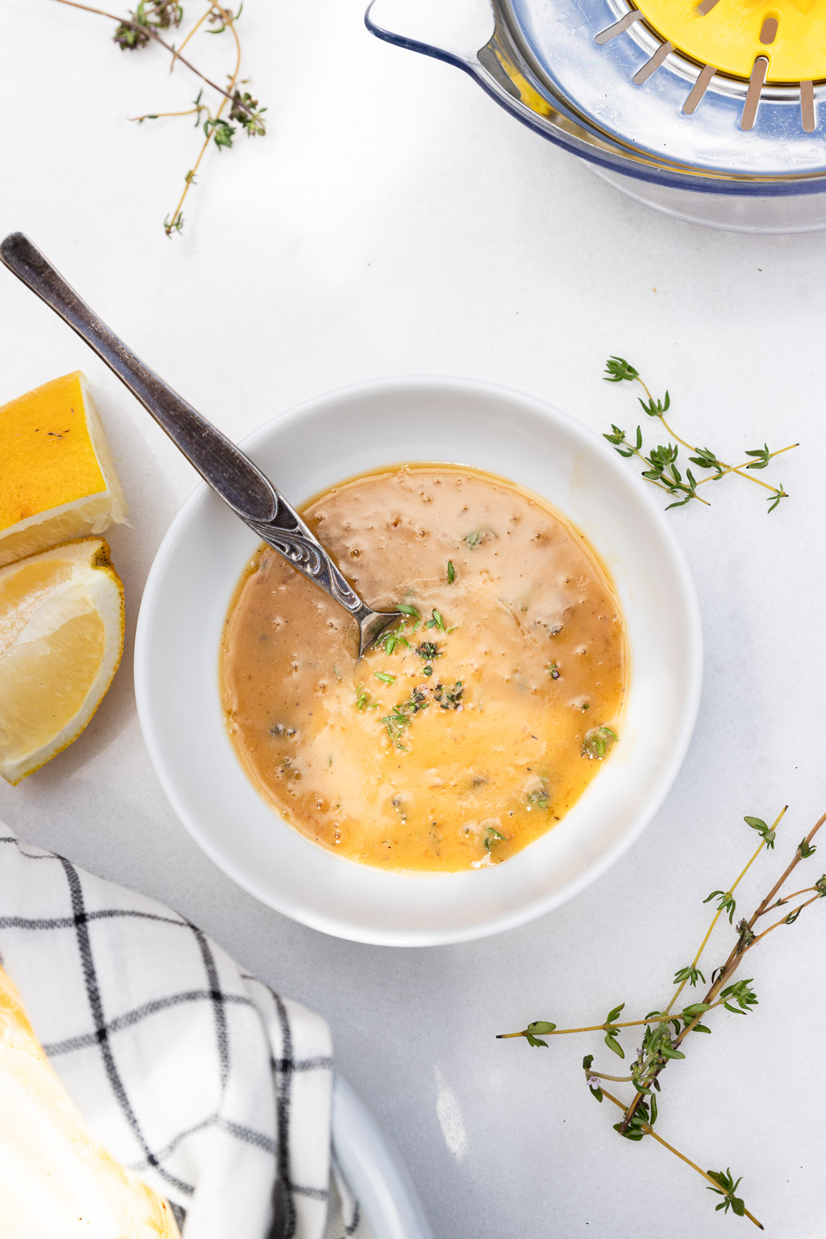 A small white dish of lemon butter sauce with a decorative spoon in it on a marble background with fresh thyme, a black and white checked tea towel and lemon at the sides.