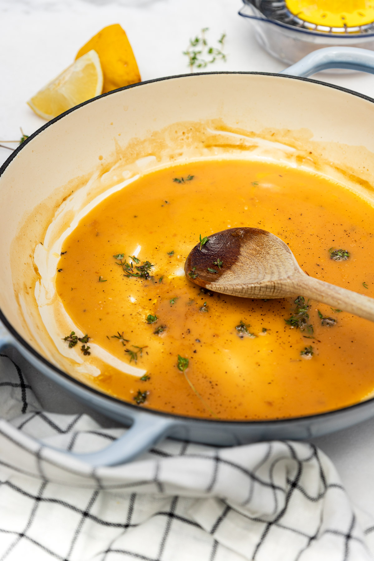 Closeup of a wooden spoon in a blue cast iron pan of lemon & butter sauce with herbs wrapped in a black and white checked tea towel and with lemon and herbs in the background.