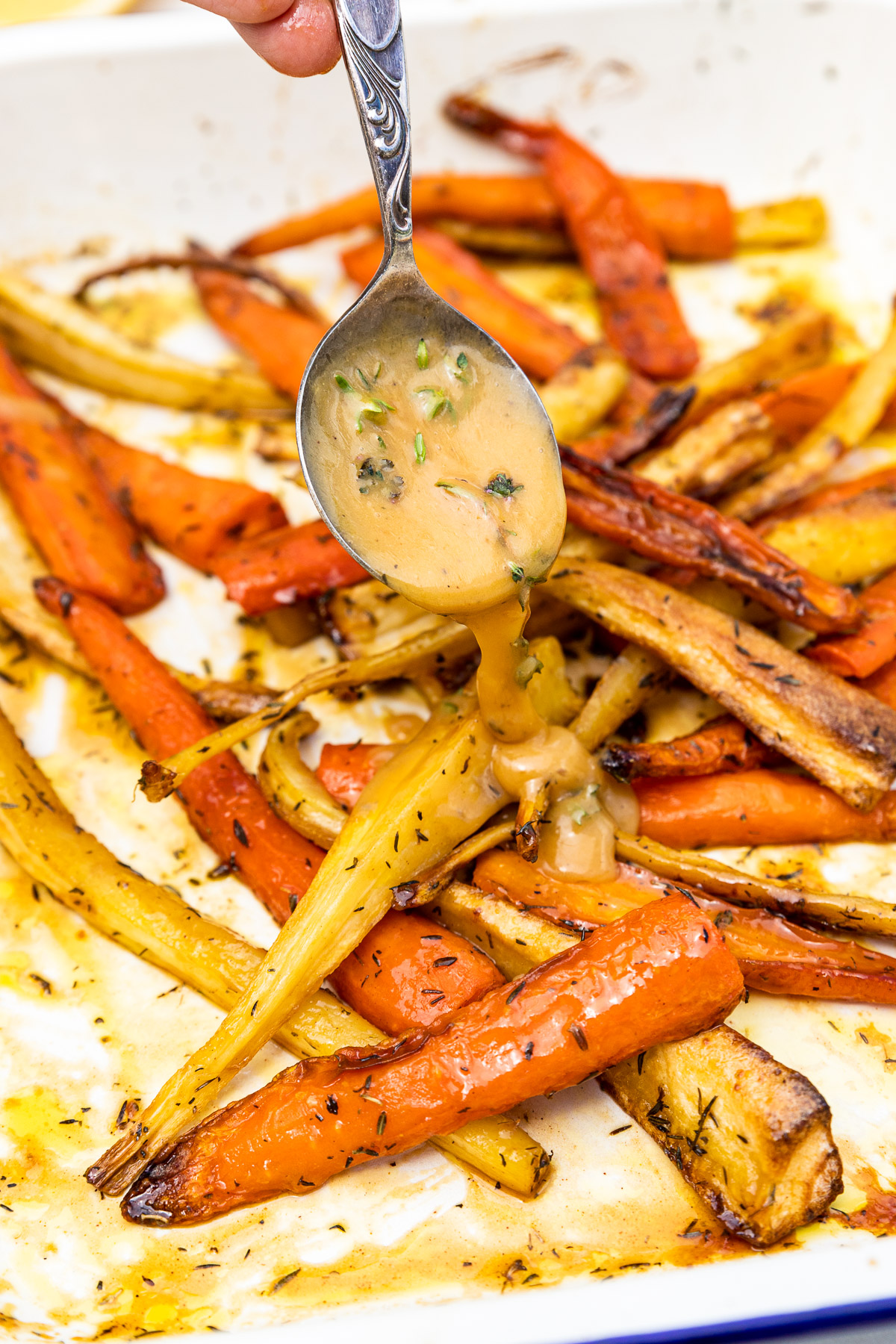 Closeup of a spoon drizzling thick lemon butter sauce with herbs over honey roast carrots and parsnips in a white baking dish with a blue rim.