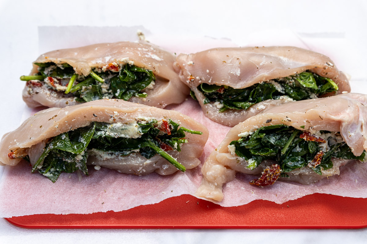 Closeup of 4 raw spinach feta stuffed chicken breasts on baking paper and a red chopping board.