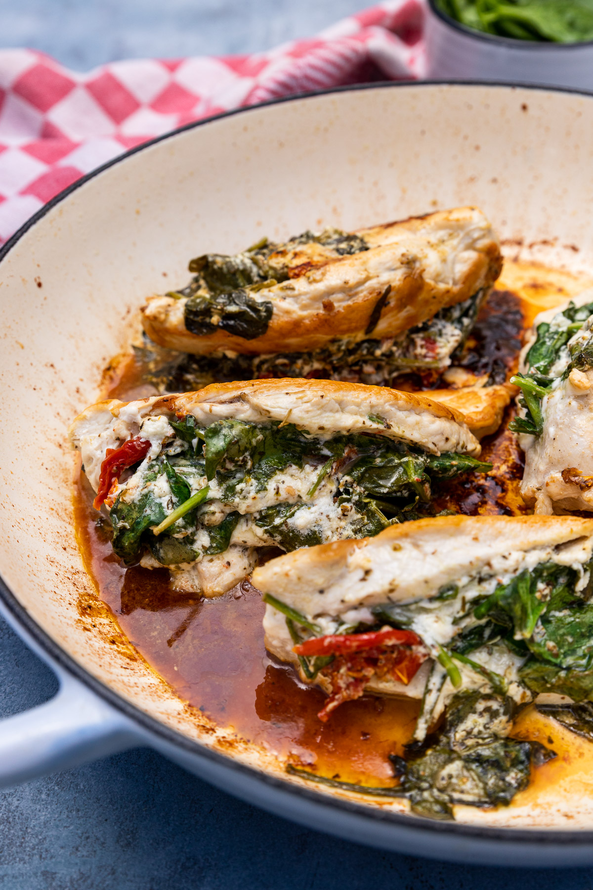 Closeup of cooked spinach feta stuffed chicken breasts in a blue cast iron pan with pan juices and on a pale blue background with a red and white checked tea towel in the background.