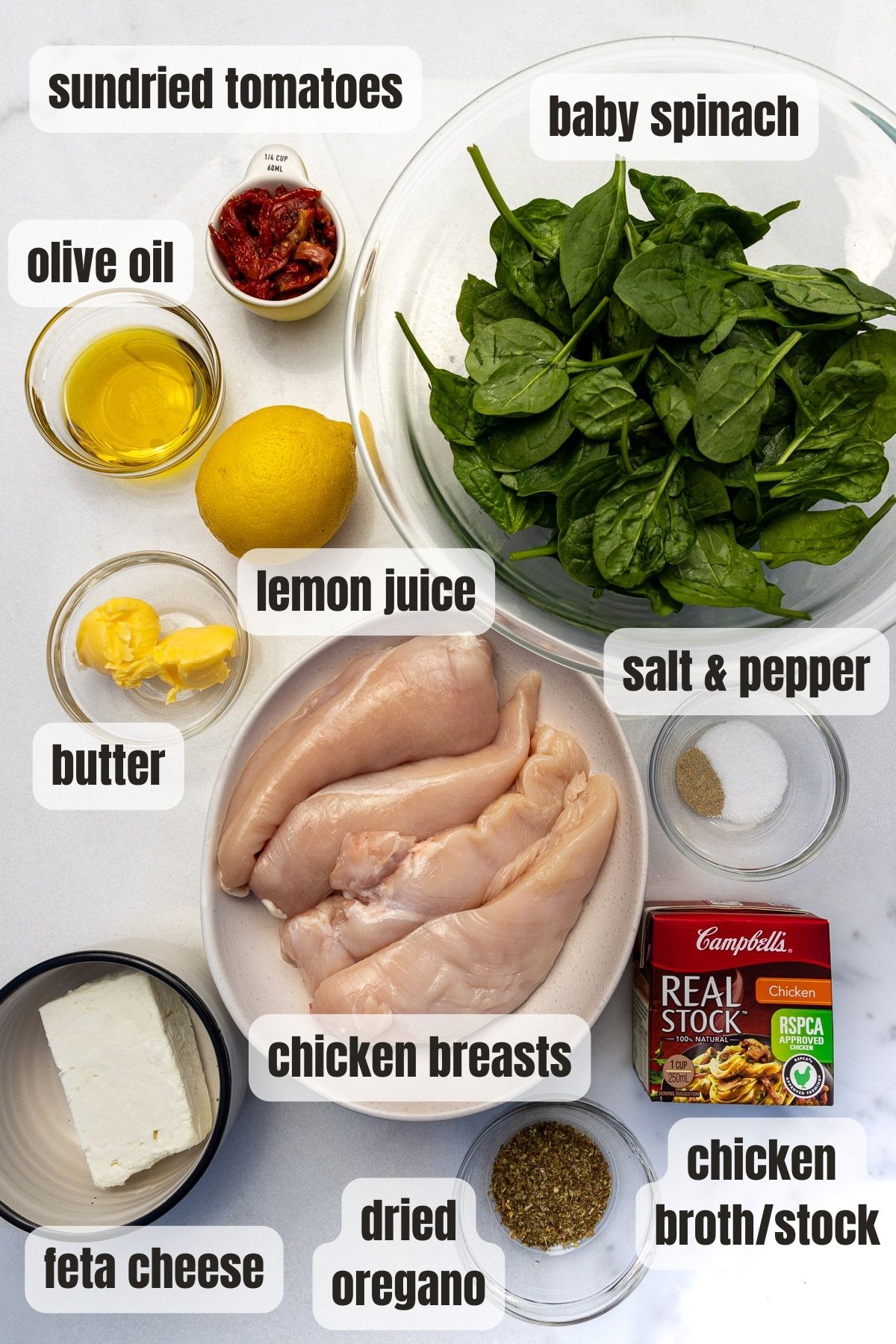 Overhead view of all the labelled ingredients for spinach feta stuffed chicken breasts including chicken breasts, baby spinach, feta, dried oregano, sundried tomatoes, olive oil, butter, lemon juice and chicken broth.