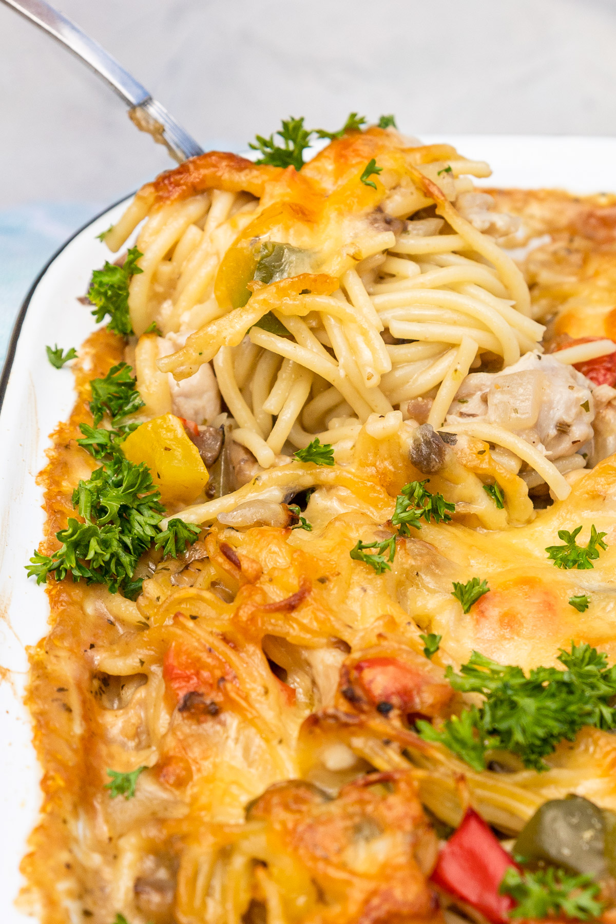 Closeup of a big serving spoon scooping out some cheesy baked chicken spaghetti from a white baking dish, all on a white background.