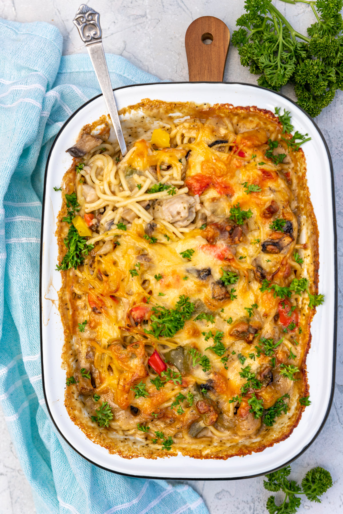 Overhead view of baked cheesy chicken spaghetti in a white dish with black rim on a grey textured background with pale blue tea towel.