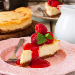 A slice of baked white chocolate cheesecake with raspberries and raspberry puree on top on a pink decorative plate with a pink tea towel and the rest of the cheesecake in the background.