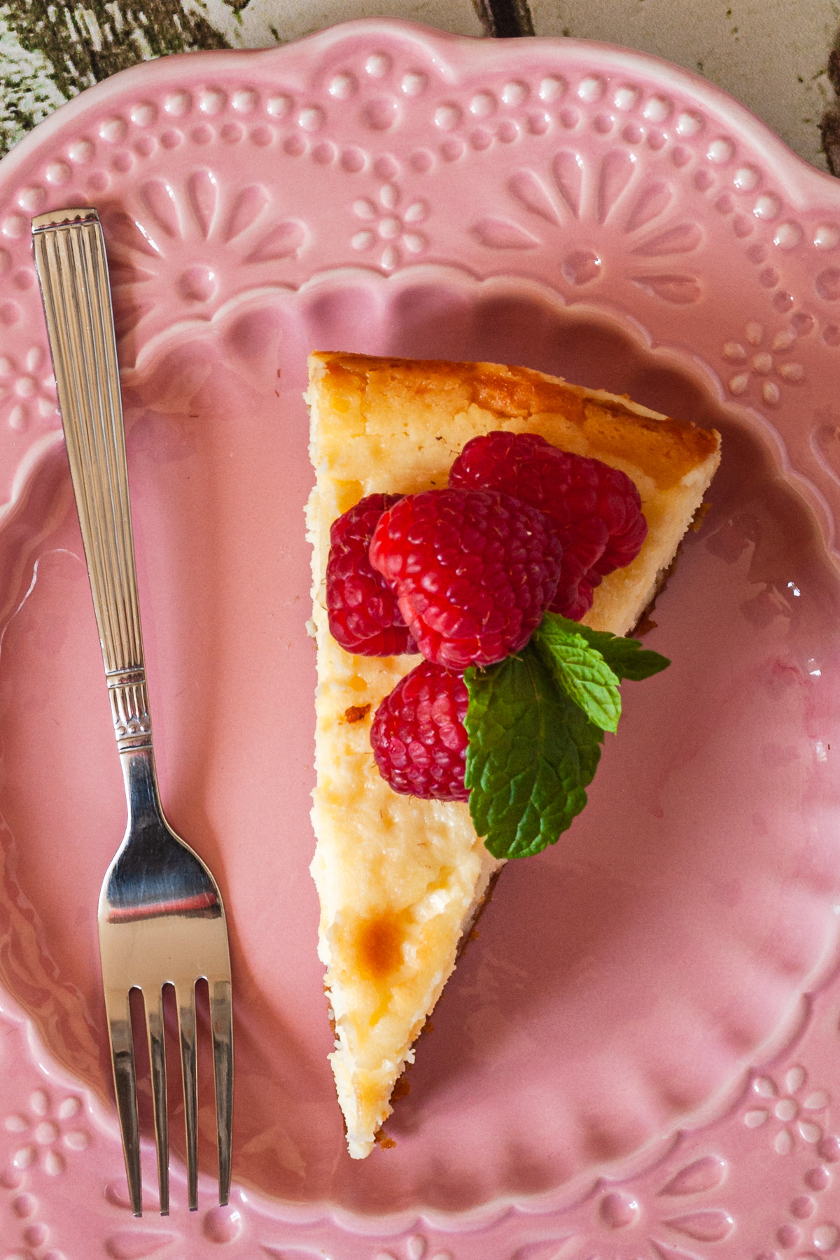 Closeup overhead view of a small slice of baked white chocolate cheesecake with raspberries and mint leaves on top, all on a decorative pink plate with a fork.