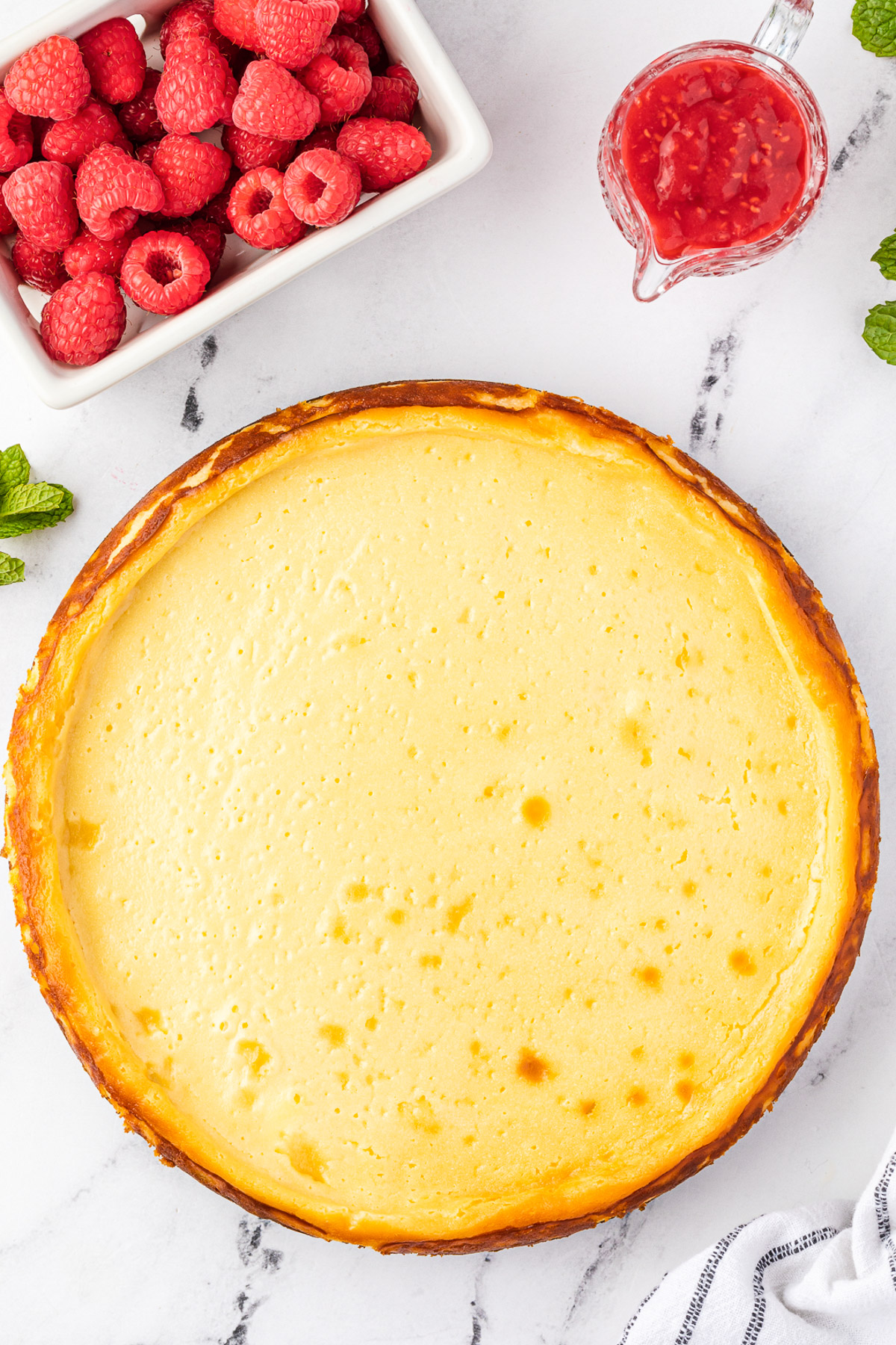 Overhead view of lightly golden baked white chocolate cheesecake just out of the oven on a marble background with raspberries and a tea towel next to it.