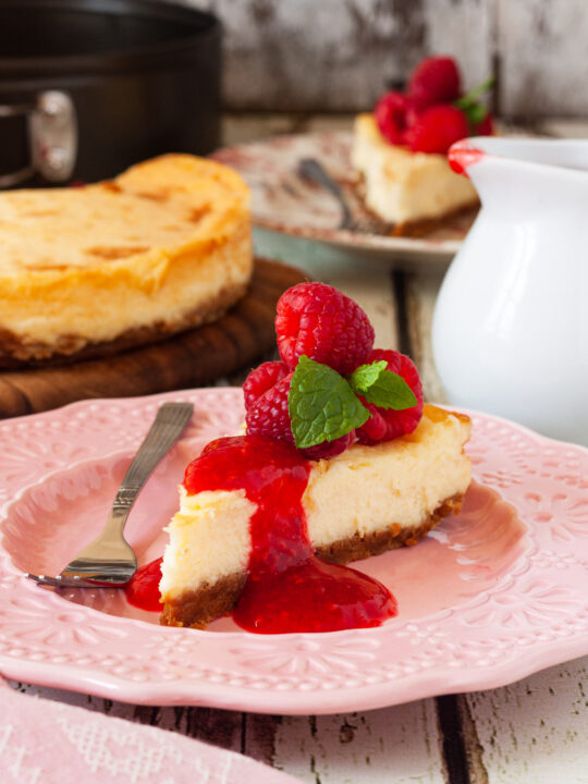 A slice of baked white chocolate cheesecake with raspberries and raspberry puree on top on a pink decorative plate with a pink tea towel and the rest of the cheesecake in the background.