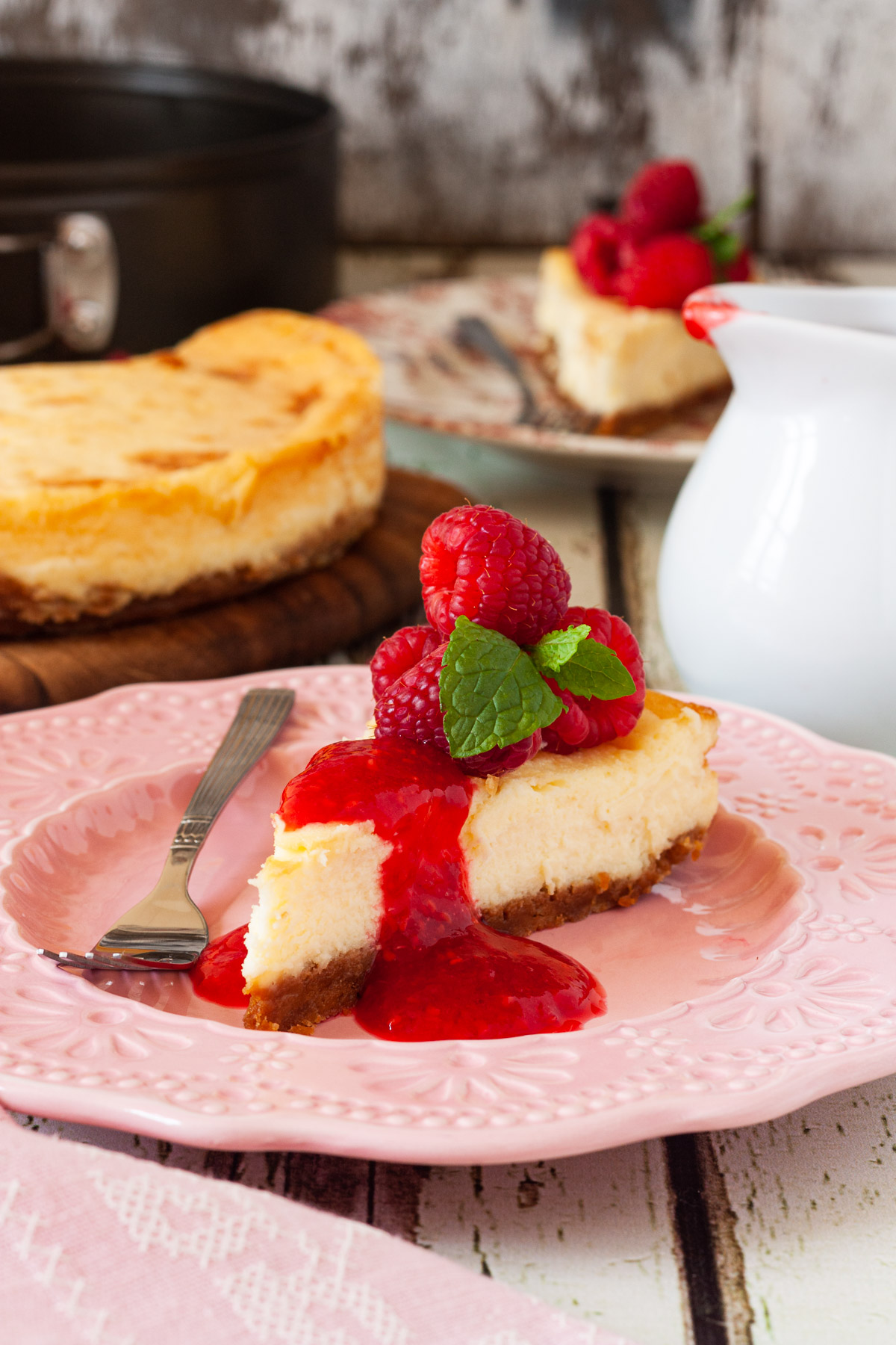 A slice of baked white chocolate cheesecake with raspberries and raspberry puree on top on a pink decorative plate with a pink tea towel and the rest of the cheesecake in the background.