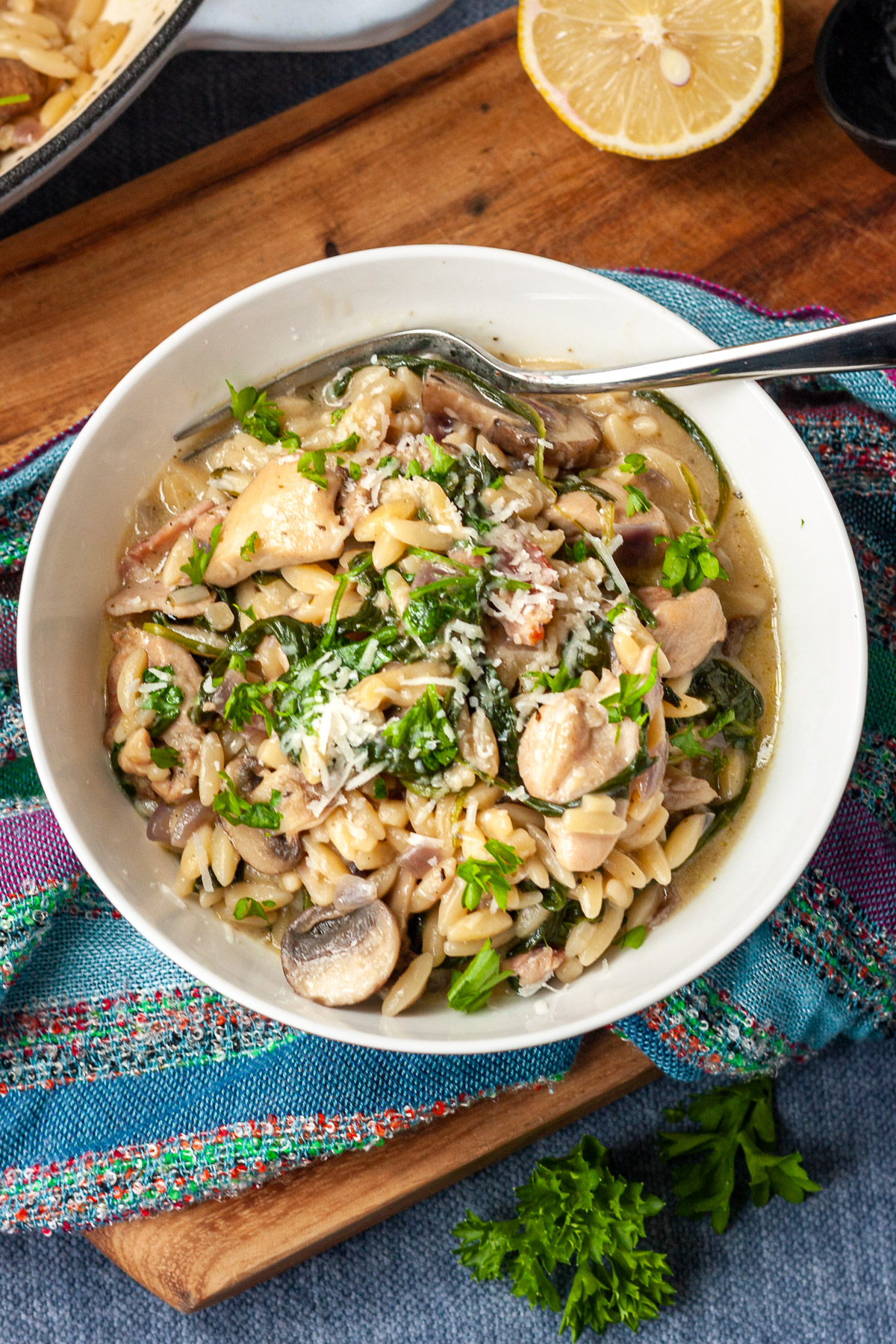 Overhead view of a bowl of creamy chicken orzo with fork in it on a multi-colored cloth and wooden board and blue background with fresh parsley and a lemon half.