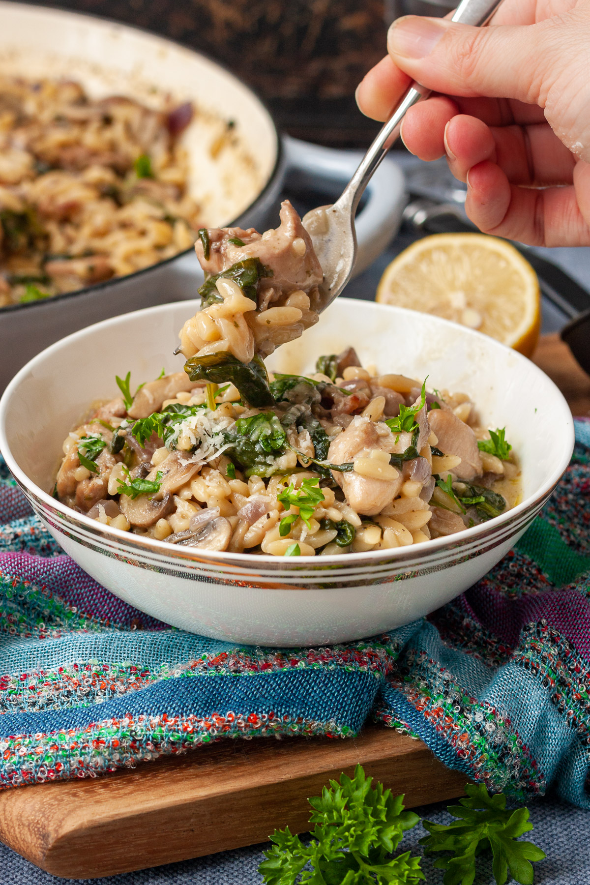 Someone eating creamy chicken orzo in a small bowl on a colorful blue and multi-colored tea towel and wooden board on a blue background with a pan and lemon in the background.