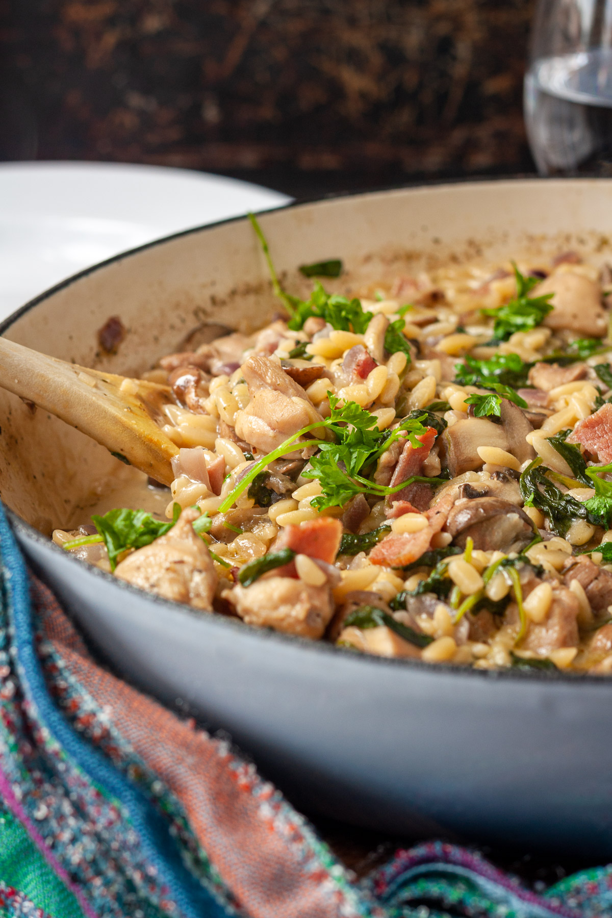 Closeup of creamy chicken orzo in half of a large blue cast iron pan with a colorful tea towel in front and a glass of water and dark background.