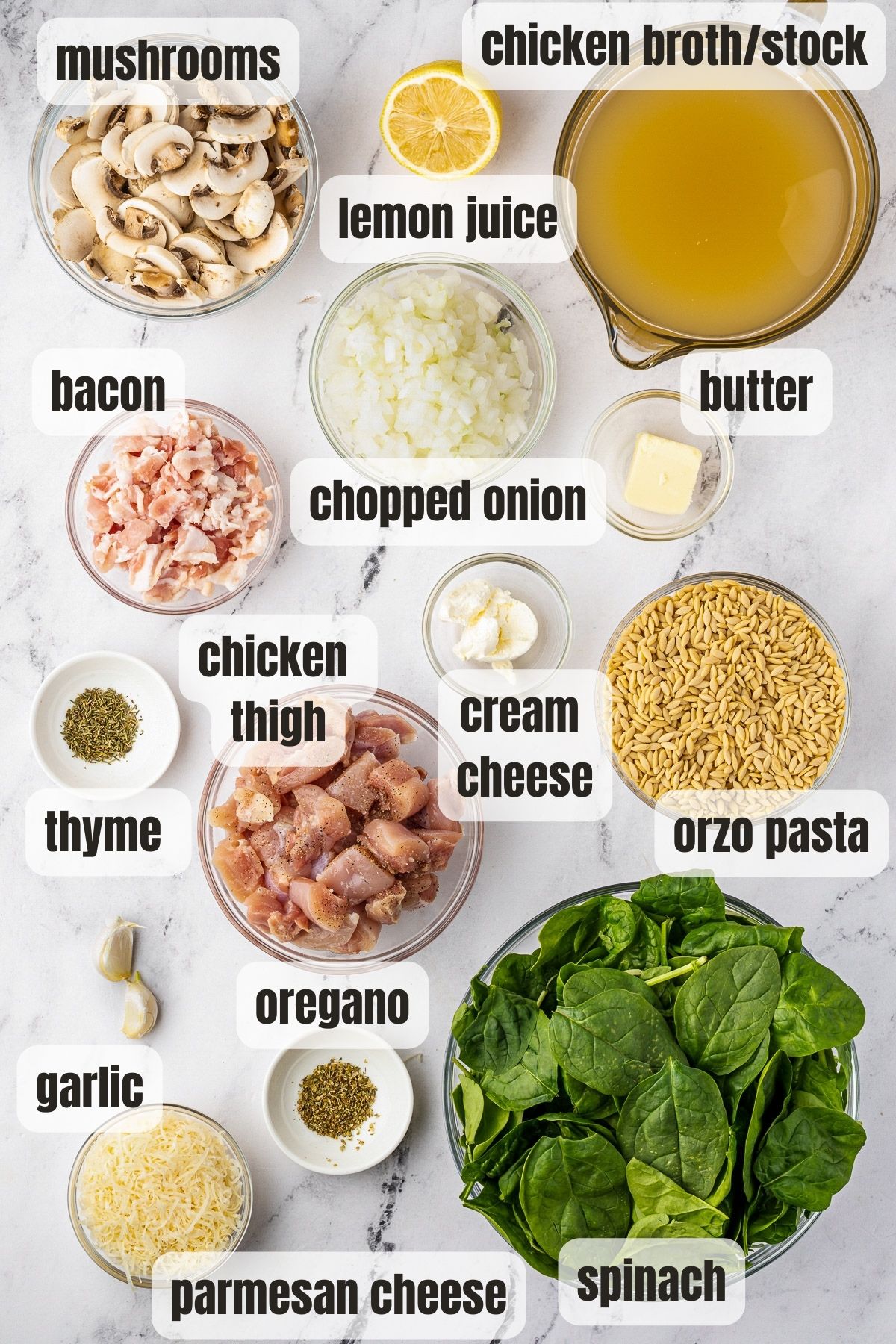 Overhead view of all ingredients needed for creamy chicken orzo including dried orzo, chopped chicken, chicken broth, onion, bacon, herbs, spinach, creamy cheese, cheese, butter, garlic and mushrooms.