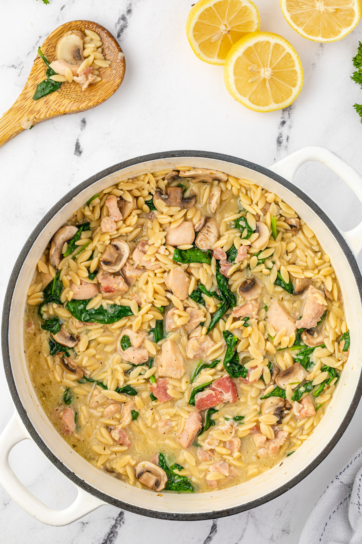Overhead view of a white cast iron pan of creamy chicken orzo on a marble background with a wooden spoon and lemon halves above.
