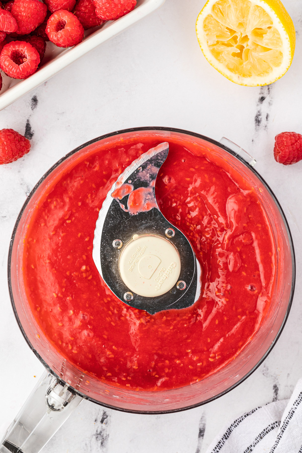 Overhead view of raspberry puree in a food processor bowl on a marble background with more raspberries and a tea towel next to it.