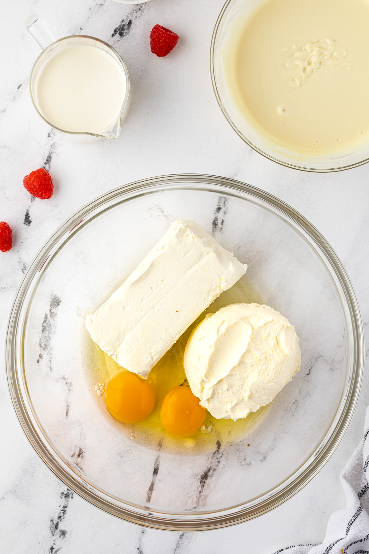 Overhead of cream cheeses and eggs in a bowl for chocolate cheesecake mixture, in a glass bowl on marble background with tea towel.