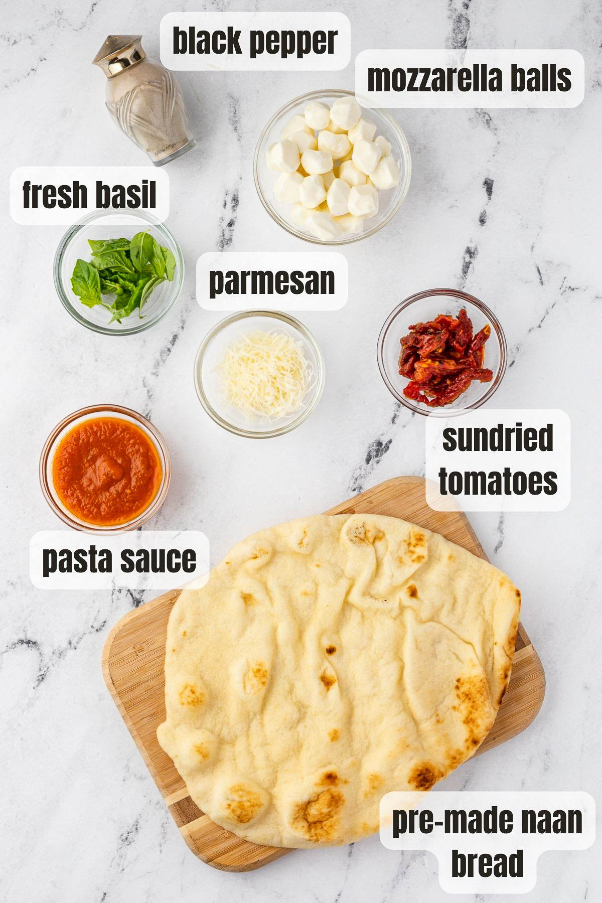 All the ingredients to make a naan pizza including pre-made naan bread, pasta sauce, sundried tomatoes, grated parmesan, fresh basil, small mozzarella balls and black pepper, all on a marble background.