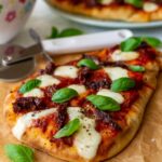 Naan pizza with mozzarella and fresh basil leaves on brown paper and a wooden board with a pizza cutter, colorful salad bowl and more pizza in the background.