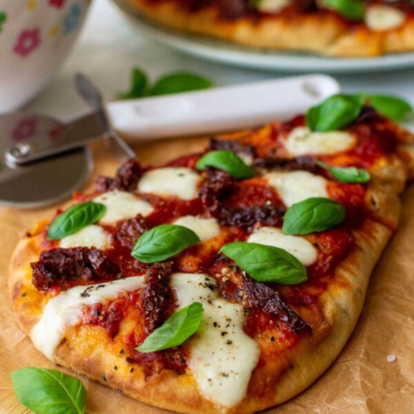 Naan pizza with mozzarella and fresh basil leaves on brown paper and a wooden board with a pizza cutter, colorful salad bowl and more pizza in the background.