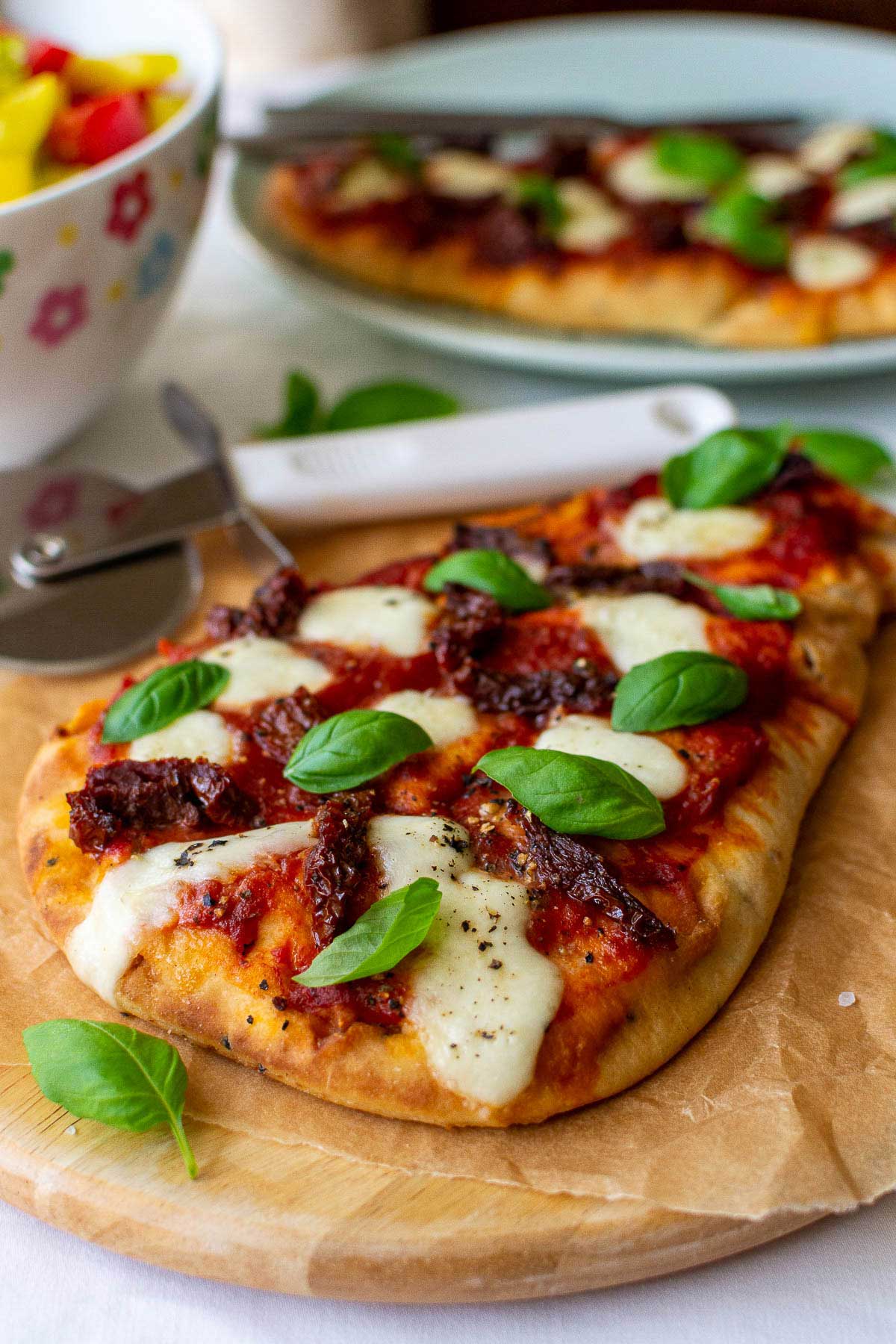 Naan pizza with mozzarella and fresh basil leaves on brown paper and a wooden board with a pizza cutter, colorful salad bowl and more pizza in the background.