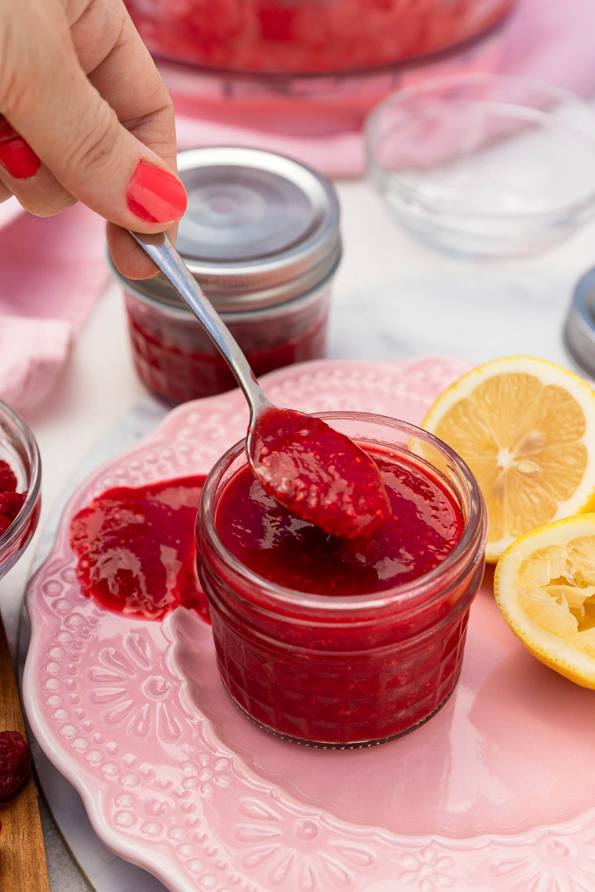 Someone spooning raspberry puree out of a small decorative glass jar on a pink decorative plate with lemons at the side and a pink tea towel in the background.