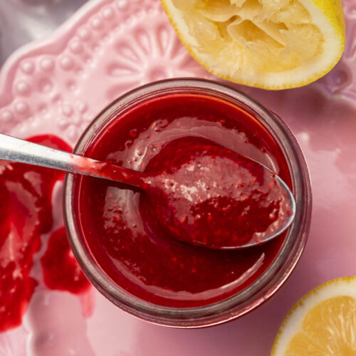 Closeup overhead of a small glass jar of homemade raspberry puree with a spoon in the top, on a pink decorative plate with lemon halves and raspberries.