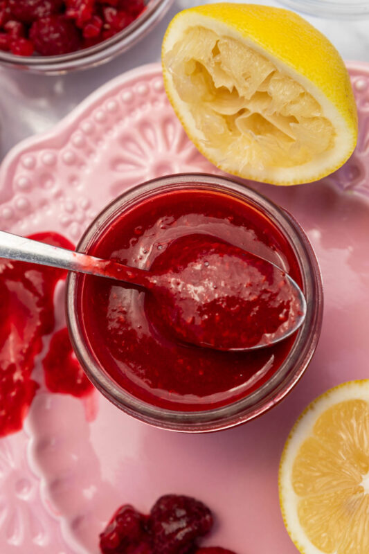 Closeup overhead of a small glass jar of homemade raspberry puree with a spoon in the top, on a pink decorative plate with lemon halves and raspberries.