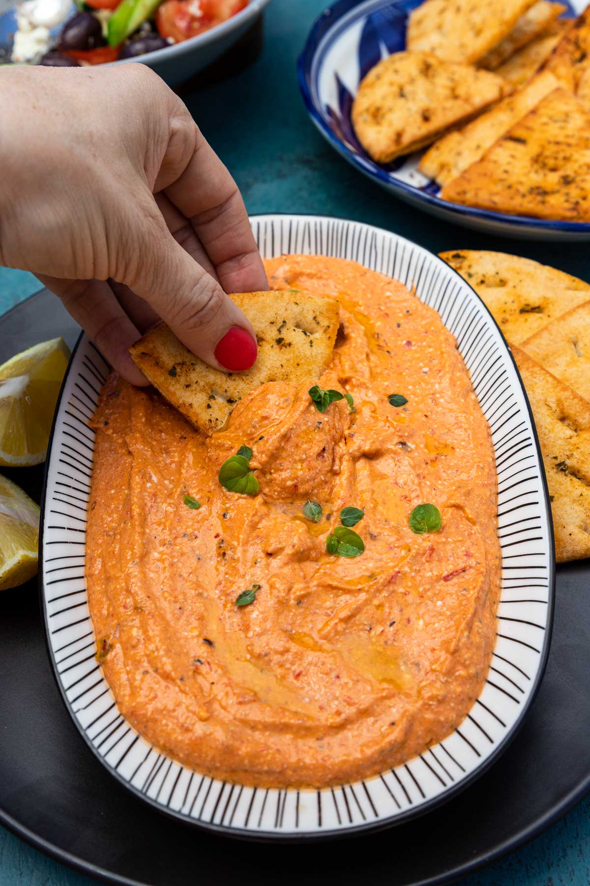 Someone scooping up spicy feta dip with a homemade pita chip from an oval shaped dish with black and white rim on a black plate and blue background with more pita chips in the background.
