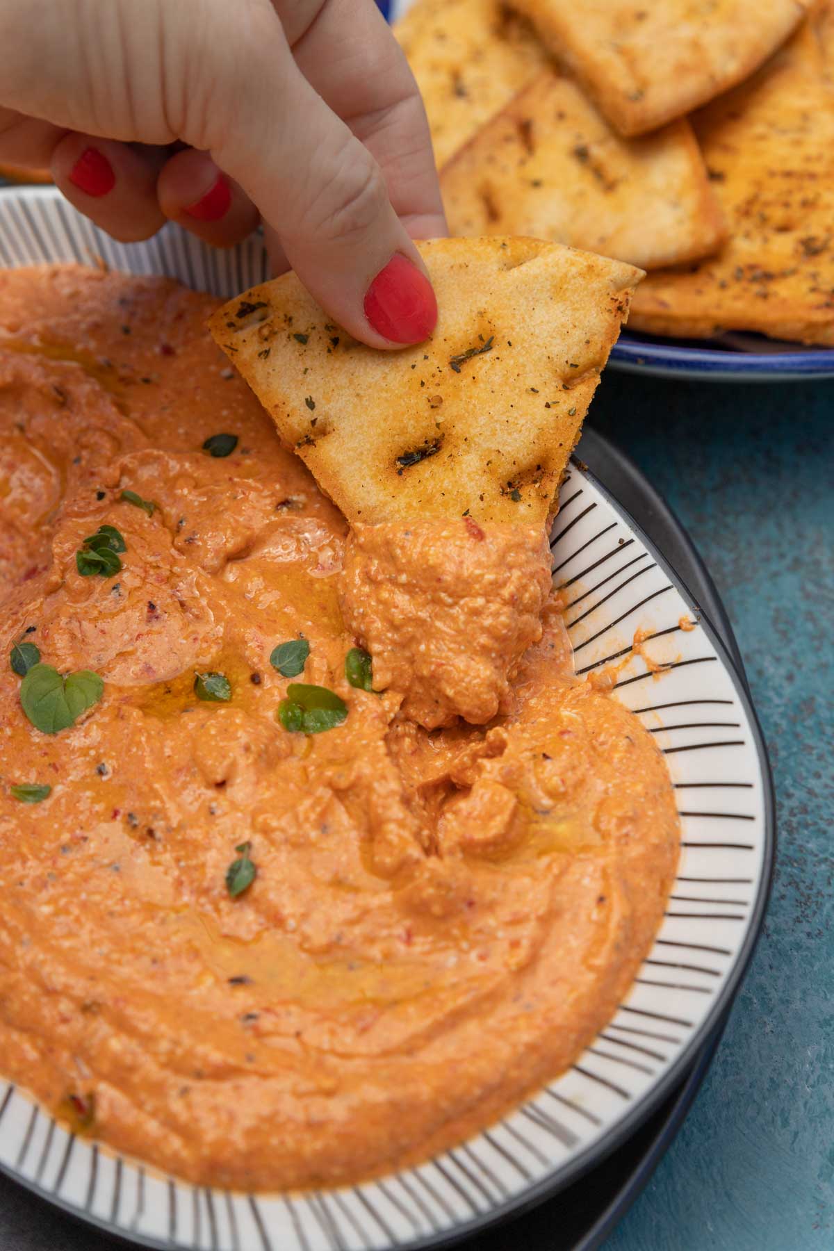 Someone scooping up some Greek spicy feta dip in an oblong shaped plate with black and white edge with a homemade pita chip, with more pita chips in the background.