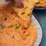 Someone scooping Greek tyrokafteri dip with a homemade pita chip from a black and white oval shaped dish on a blue background.