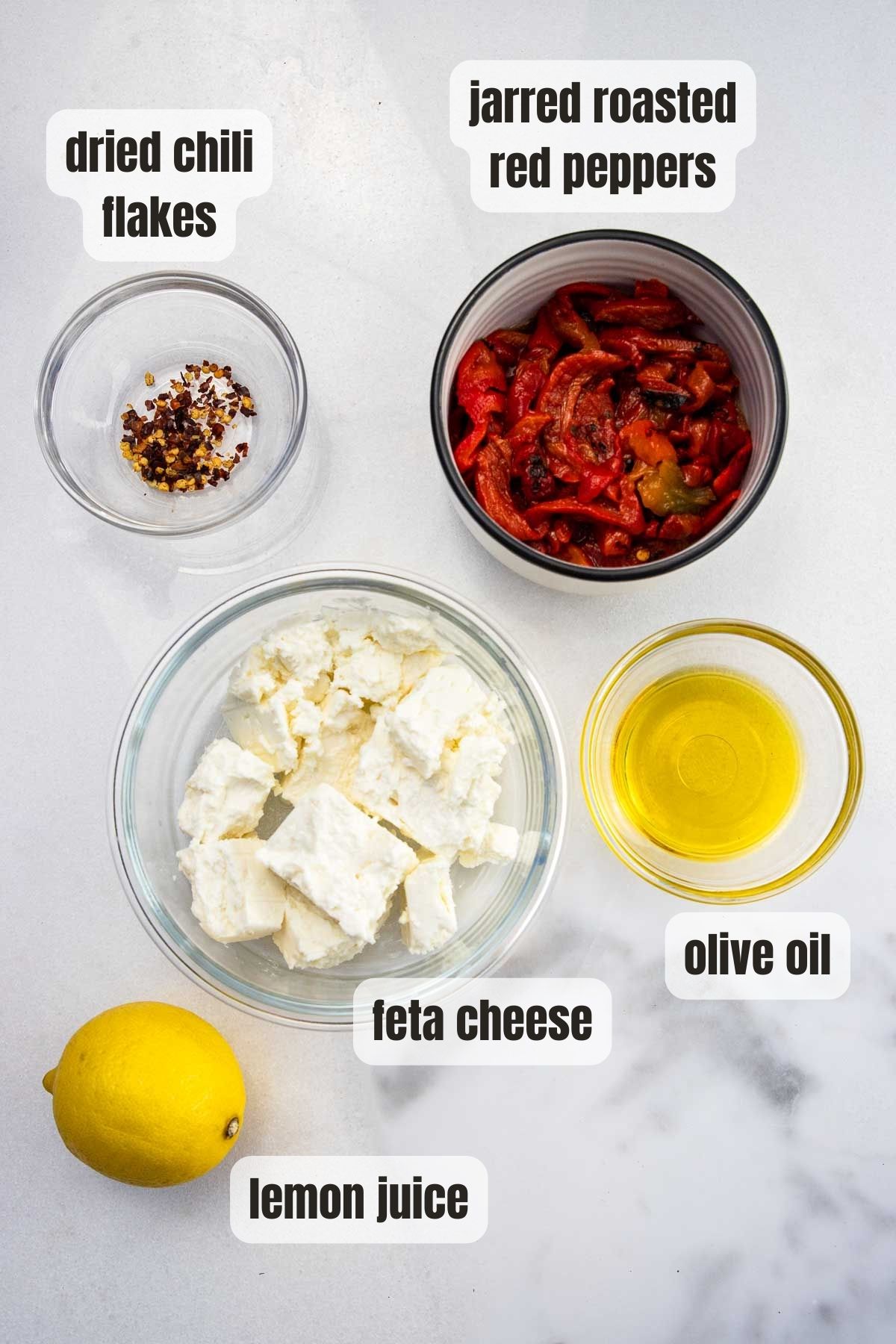 Overhead of all the ingredients needed to make tyrokafteri or spicy feta dip including feta cheese, jarred red peppers, lemon juice, olive oil and dried chili flakes.