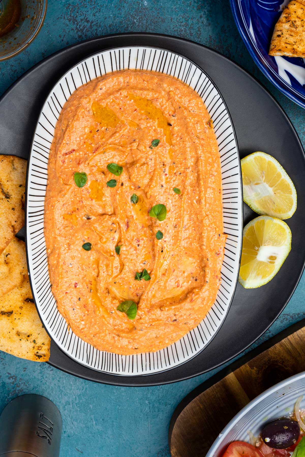 A black and white oblong shaped dish of spicy Greek feta dip with a bit of fresh oregano on top and with salt, Greek salad, pita chips and lemon halves next to it, all on a blue background.