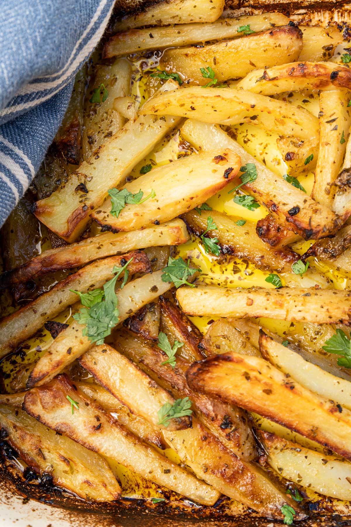 A closeup of an overhead of some golden brown cooked Greek potatoes in a baking dish with a blue tea towel in the corner and with a sprinkle of parsley on top.