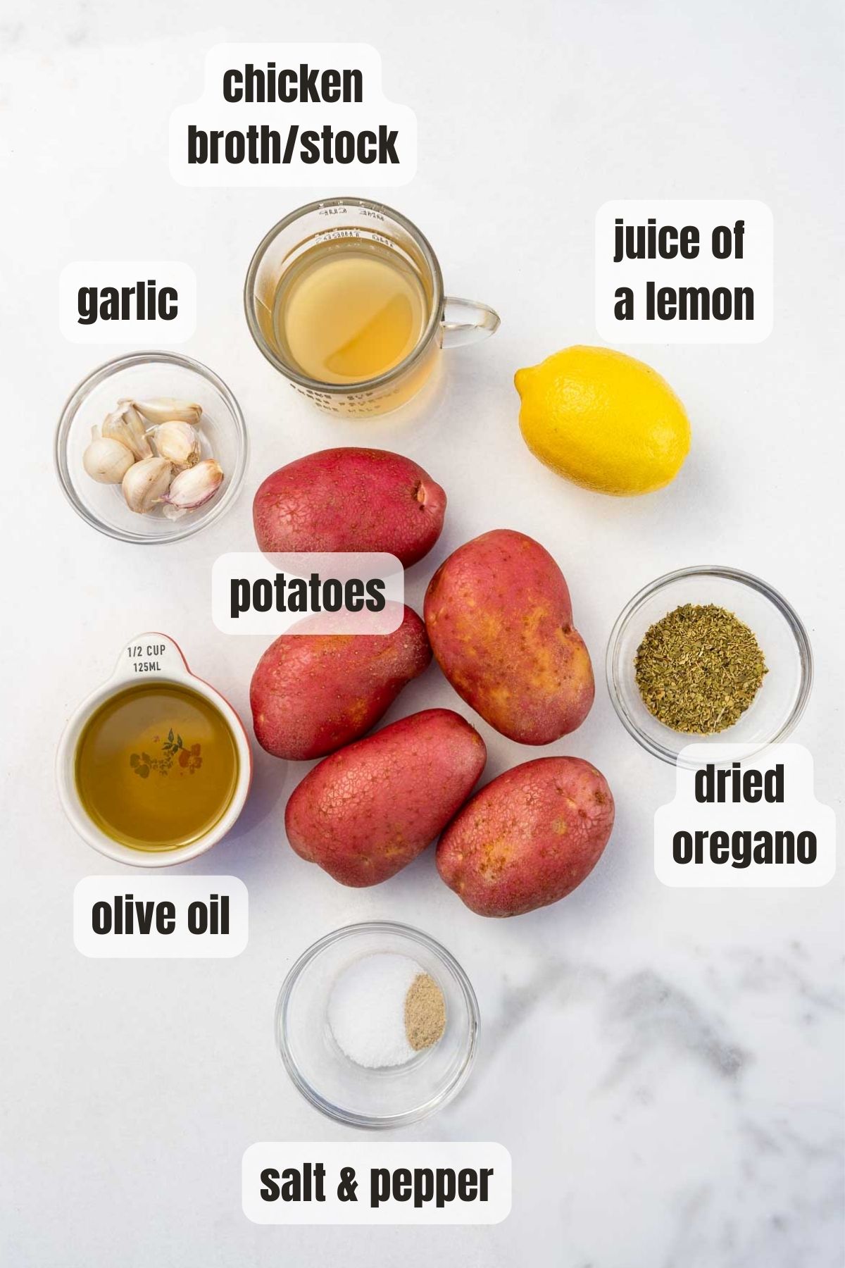 Overhead of all the ingredients needed to make Greek potatoes including potatoes, olive oil, lemon juice, dried oregano, garlic cloves, salt and pepper and chicken broth, all on a marble background.