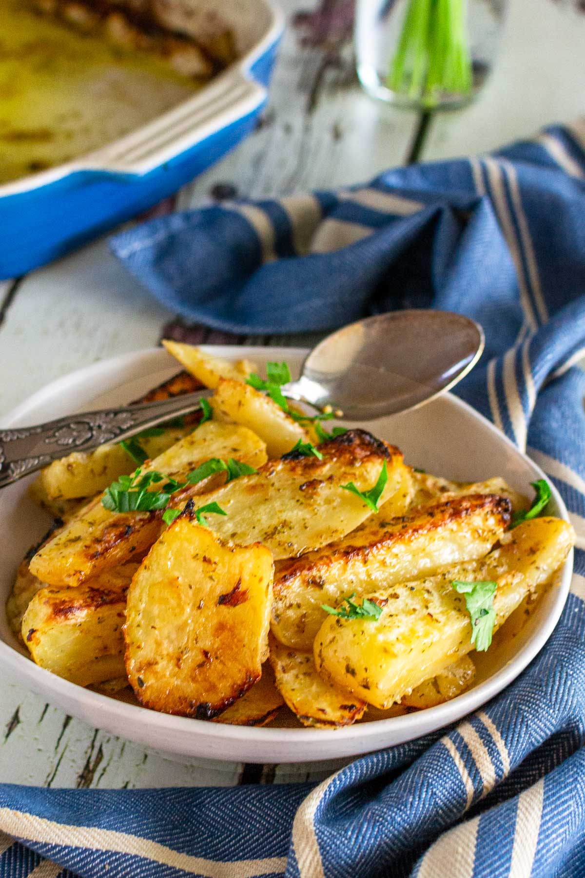 A small dish of Greek potatoes in a beige dish with a spoon in and parsley on top, with a blue and white striped tea towel in front and a blue baking dish at the back, all on a white wooden background.