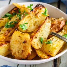 A closeup of a dish of cooked Greek potatoes with a spoon in the dish and a sprinkle of parsley on top, with a blue and white striped tea towel in front.