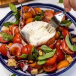 Someone's hands shown cutting into a whole burrata cheese on top of a caprese style salad with cherry tomatoes, red onion, croutons and fresh basil, in a blue bowl on a marble background.