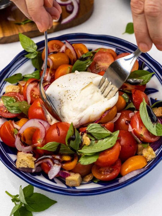 Someone's hands shown cutting into a whole burrata cheese on top of a caprese style salad with cherry tomatoes, red onion, croutons and fresh basil, in a blue bowl on a marble background.