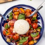 An overhead view of a burrata caprese salad with cherry tomatoes, red onion, croutons and fresh basil in a blue patterned blow with a big whole ball of burrata cheese in the middle, and with serving spoons in it.