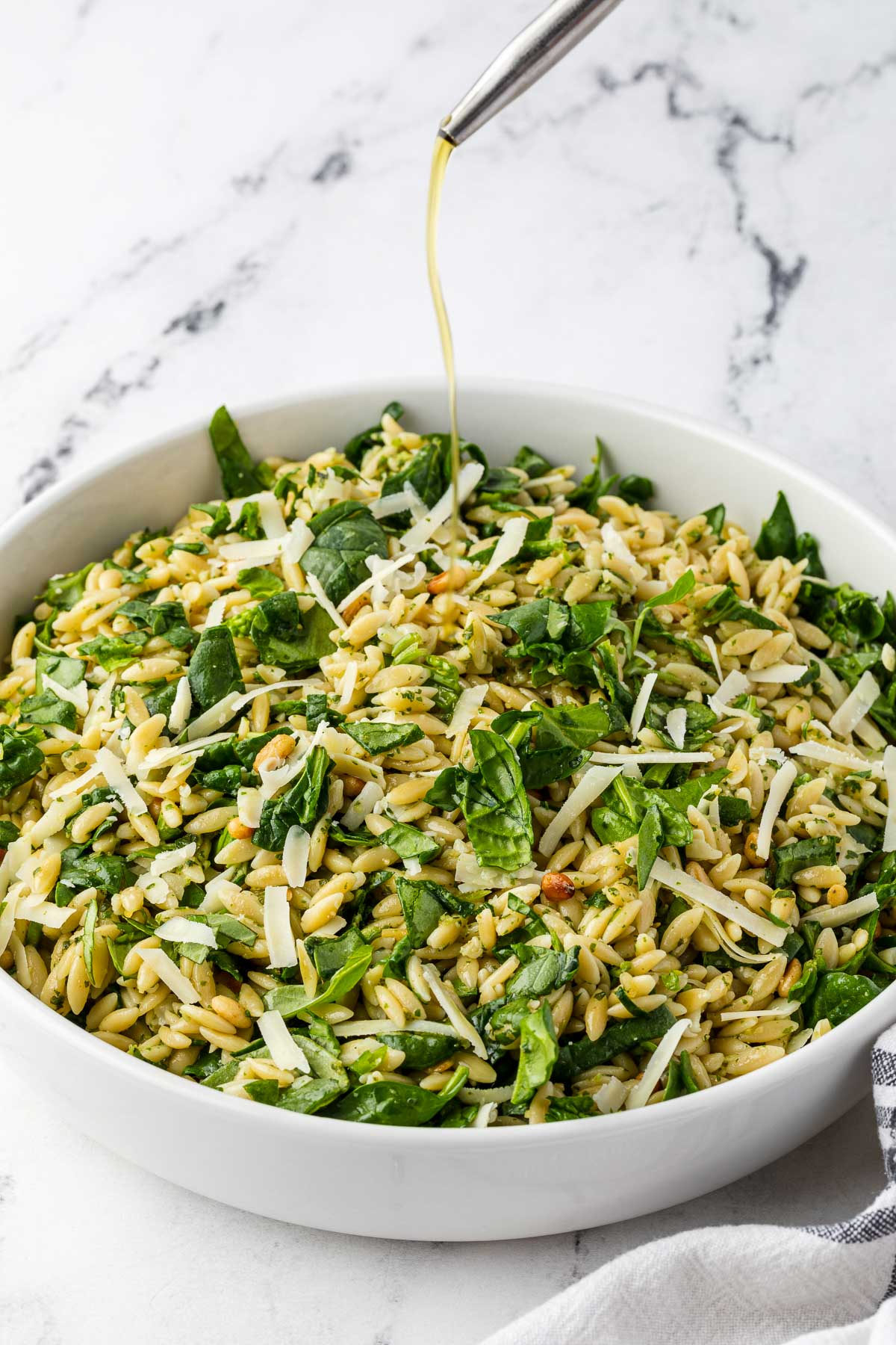 A closeup of a white bowl of pesto orzo salad with chopped spinach, parmesan and pine nuts visible on the top, and with someone drizzling olive oil over the bowl.