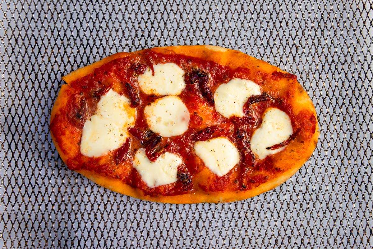 Overhead view of a small pizza in a large air fryer basket with mozzarella cheese and sundried tomatoes on top.