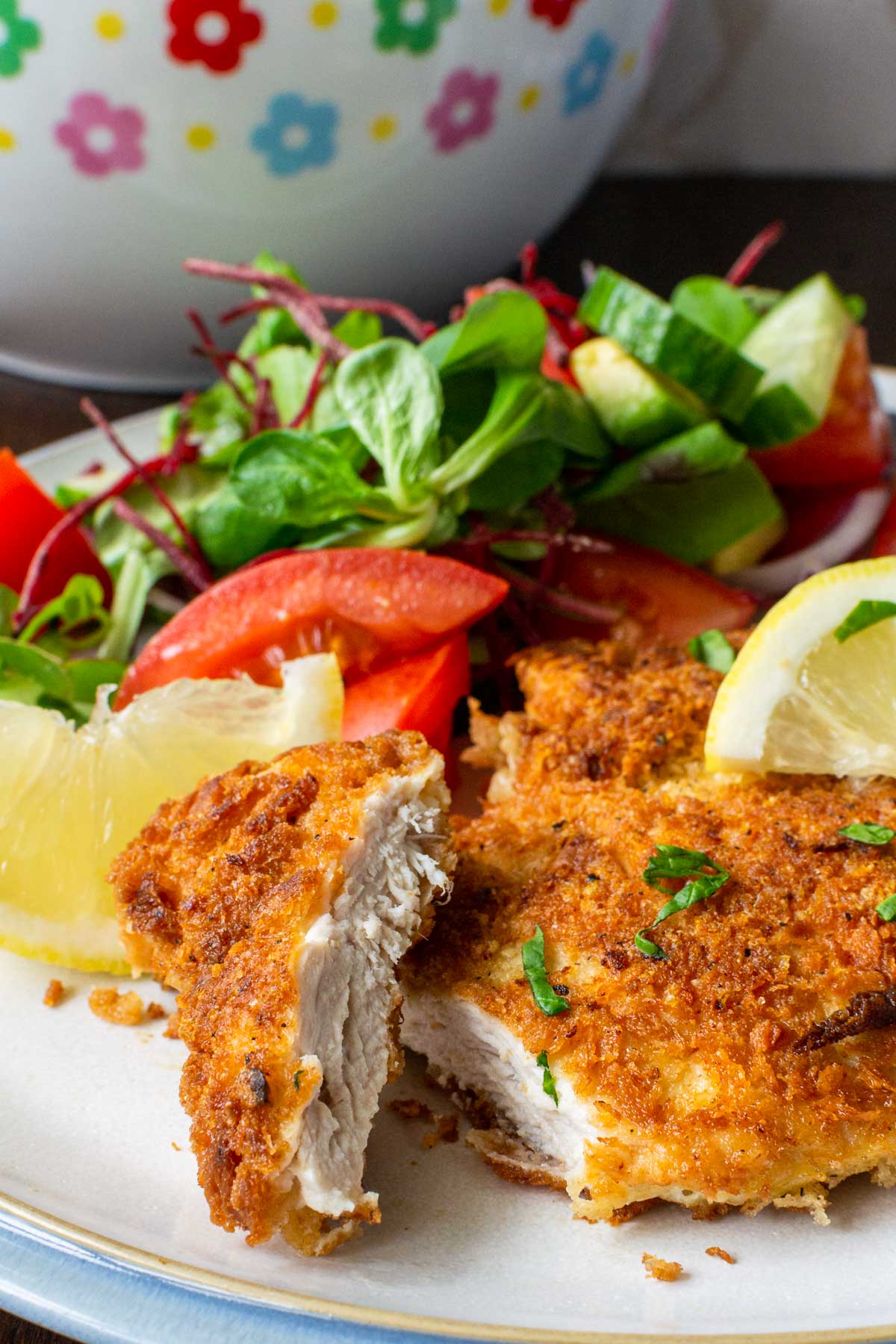 Closeup view of the inside of a piece of panko chicken breast on a white plate with blue rim with a simple salad on the plate in the background, plus salad bowl.